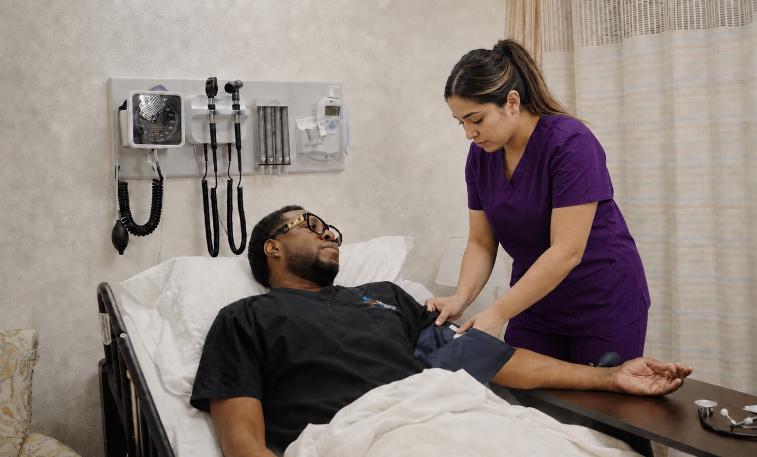 Adult CNA student in purple scrubs practicing a blood-pressure check in a training laboratory